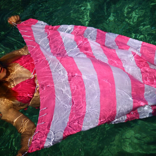A lady floats the Hammamas bold watermelon white Turkish towel in the pool.